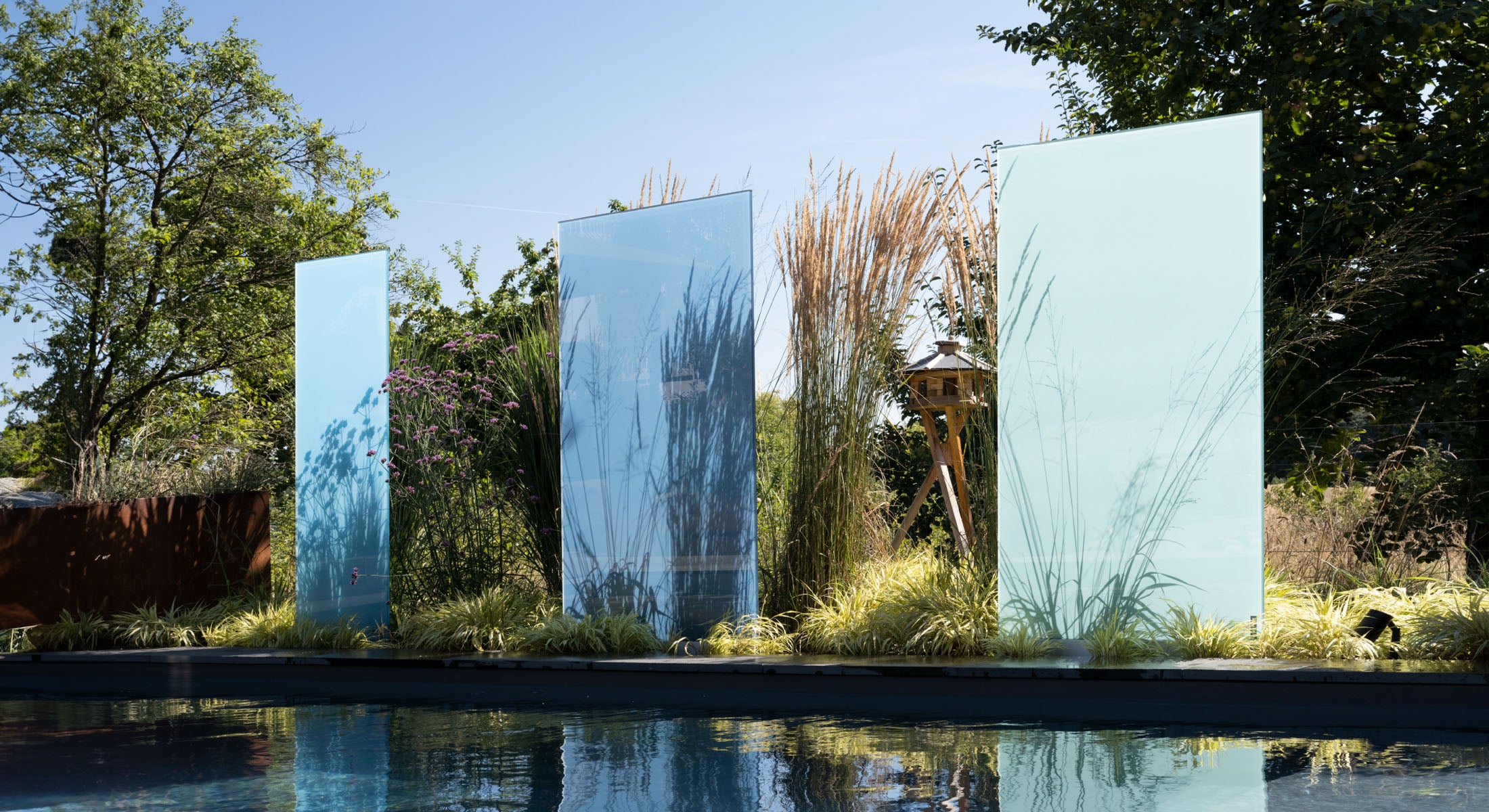 Blaue KNUMOX-Glasdesignstelen, die in ihrer kunstvollen Gestaltung und leuchtenden Farbgebung faszinieren, stehen an einem idyllischen Naturteich, der von einer üppigen Vielfalt an Pflanzen umgeben ist. Diese Pflanzen schaffen nicht nur eine harmonische Verbindung zur Natur, sondern tragen auch zu einem reizvollen Schattenspiel auf den glatten, glänzenden Scheiben der Stelen bei, das sich im Laufe des Tages mit dem Sonnenlicht verändert und so ein dynamisches visuelles Erlebnis bietet.