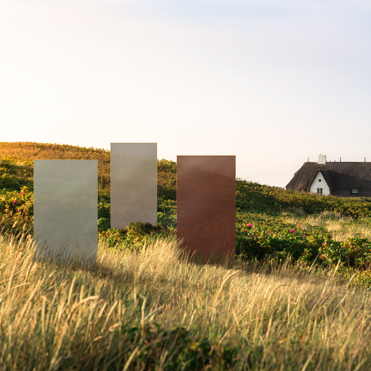Das KNUMOX Element Dolomite mit beiger Farbe steht mit 2 anderen Glaselementen auf einem hügeligen Feld, Im Hintergrund ist ein Haus mit Reetdach zu sehen.