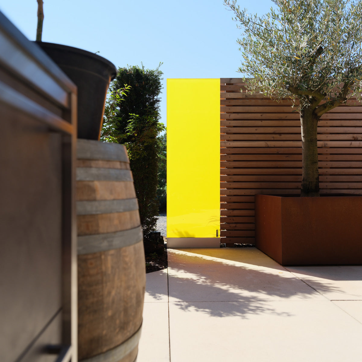 Das KNUMOX Glasdesign-Element Honey mit leuchtend gelber Farbe steht in einem Garten zwischen einer Hecke und einem Holzzaun.