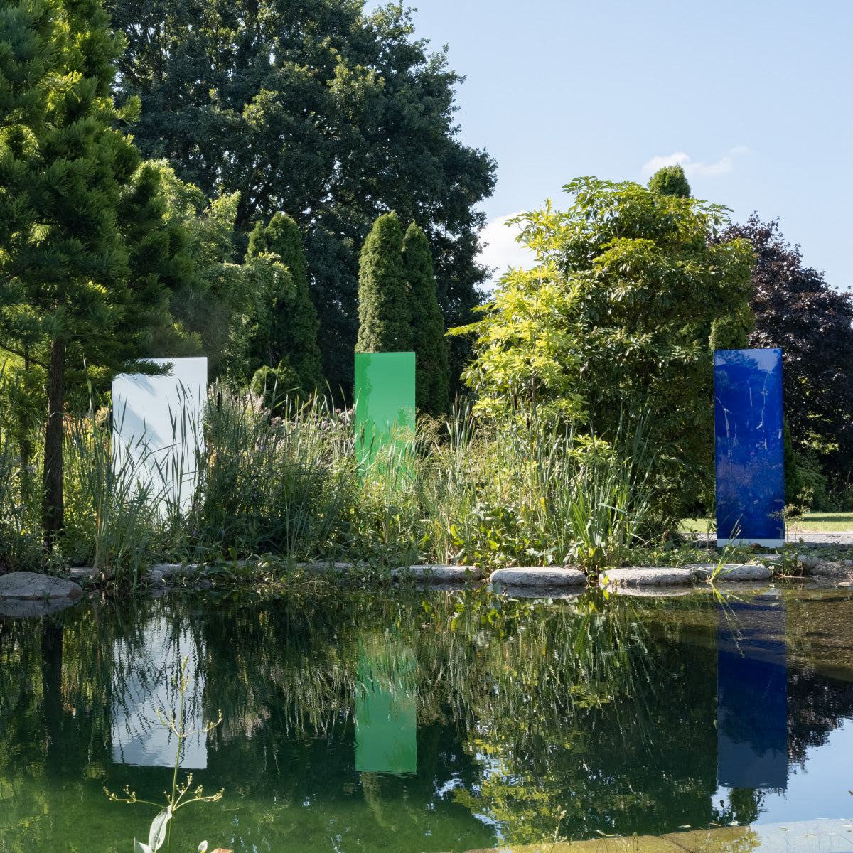 Eine KNUMOX Kollektion miteinem weißen, einem grünen und einem blauen Glaselement stehen in einem Garten in üppiger Bepflanzung vor einem See.