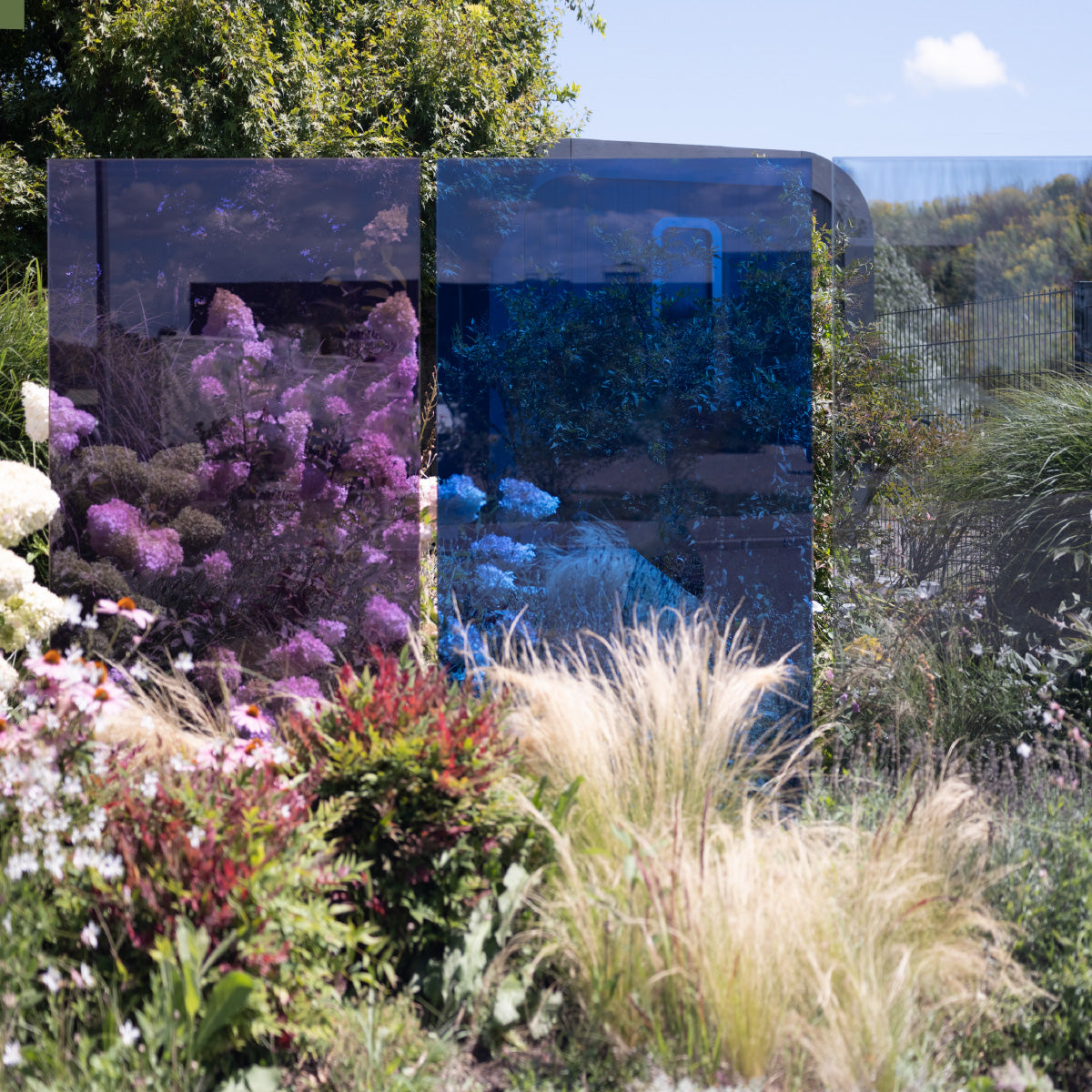 Eine violette und eine blaue Glasdesign Stele stehen in der Pflanzung, die Elemente sind durchlässig so dass man auch die Pflanzen dahinter sehen kann.