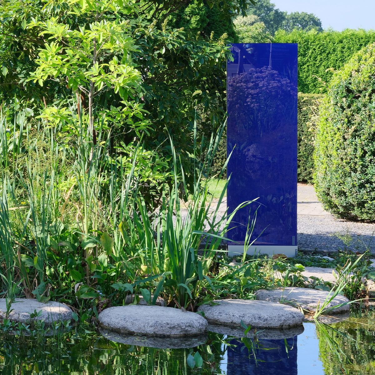 Blaues Glaselement steht zwischen Pflanzen an einem Teich mit Spiegelung im Wasser.